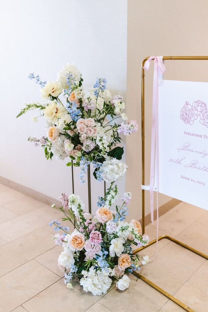 pastel floral tower next to a custom event sign with hand-drawn peonies mounted on a gold easel with draping pink silk ribbons
